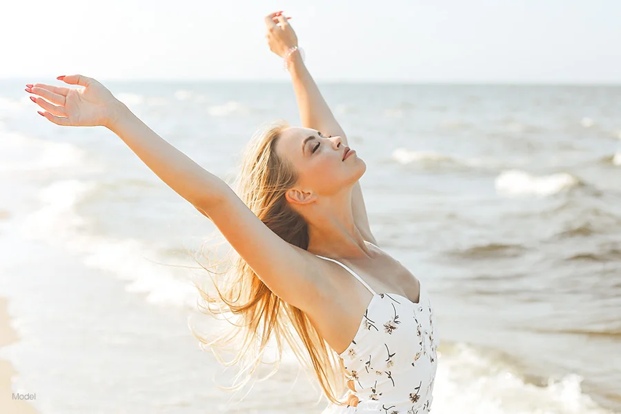 A beautiful blonde woman in a white floral sundress standing on a beach with her arms raised, facing the sunlight and ocean waves.