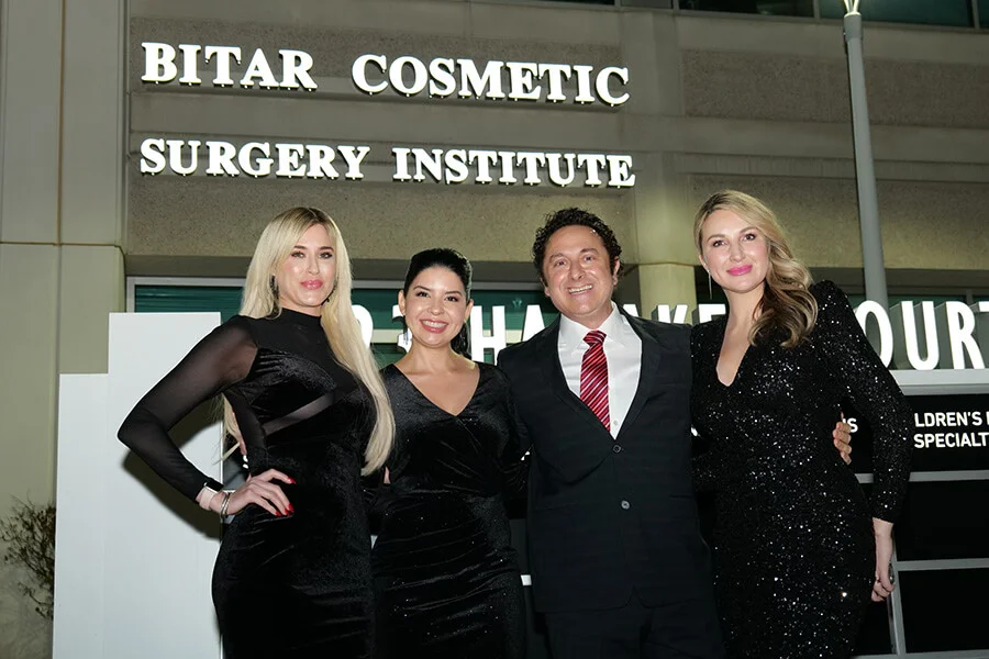 Dr. Bitar and three female team members, all wearing black evening attire, stand smiling beneath the illuminated sign of the Bitar Cosmetic Surgery Institute at night.