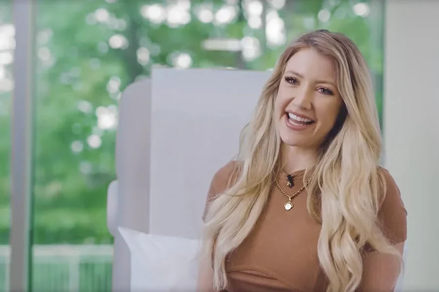 A smiling, happy blonde woman with long wavy hair, wearing a brown top and gold necklaces, sitting in a light, bright setting with greenery visible in the background.