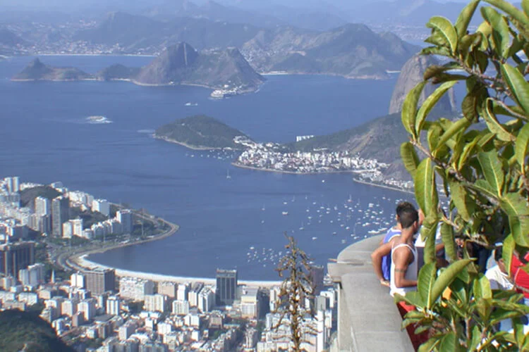 An aerial view overlooking Rio de Janeiro, showing the curved Copacabana beach, Ipanema, Sugarloaf Mountain, Guanabara Bay, and surrounding mountains, with people observing the scene in the foreground.