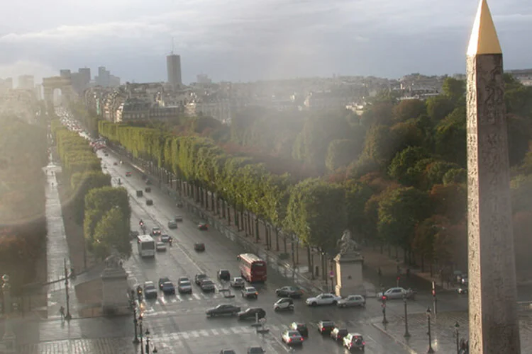 An aerial view of the Avenue des Champs-Élysées in Paris, with traffic on a wet street, lined with trees, leading toward the Arc de Triomphe, with the Luxor Obelisk in the foreground.