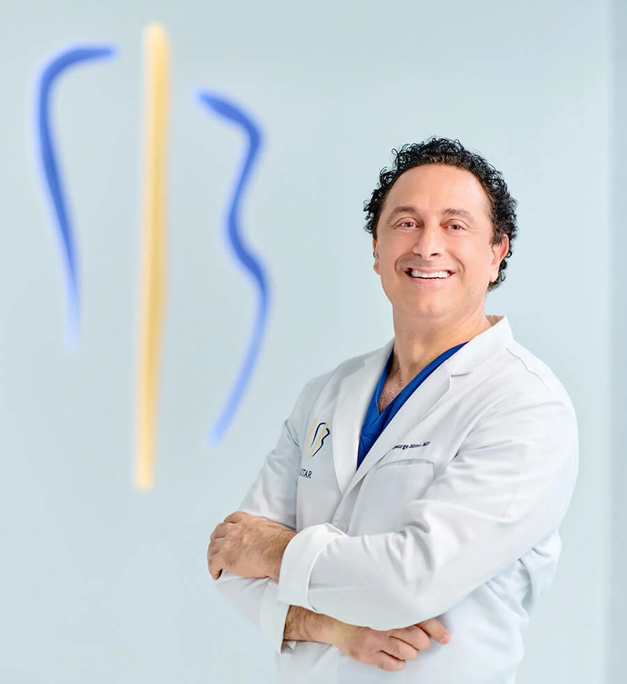A headshot of a smiling Dr. Bitar in a white lab coat with his arms crossed, standing in front of a wall featuring a soft blue and yellow neon logo.