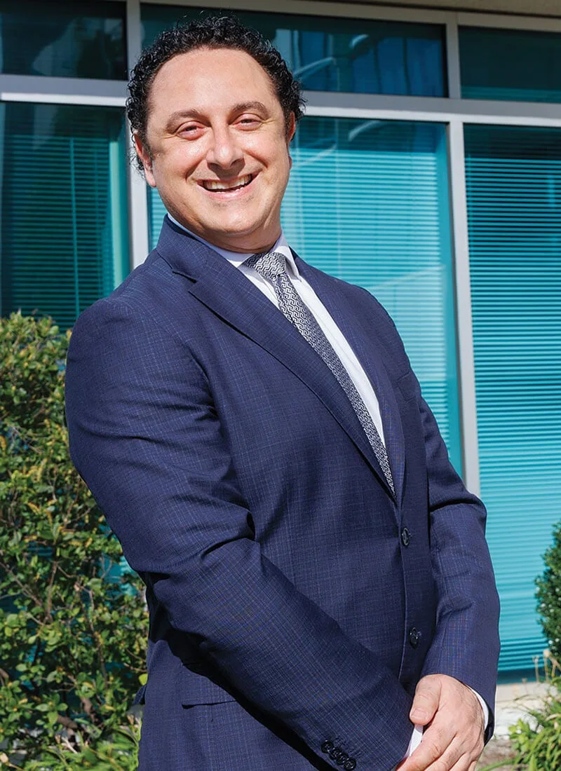 A photo of a smiling Dr. Bitar, a man with dark curly hair, wearing a dark blue suit and a patterned tie, standing outdoors in front of blue glass windows and greenery.