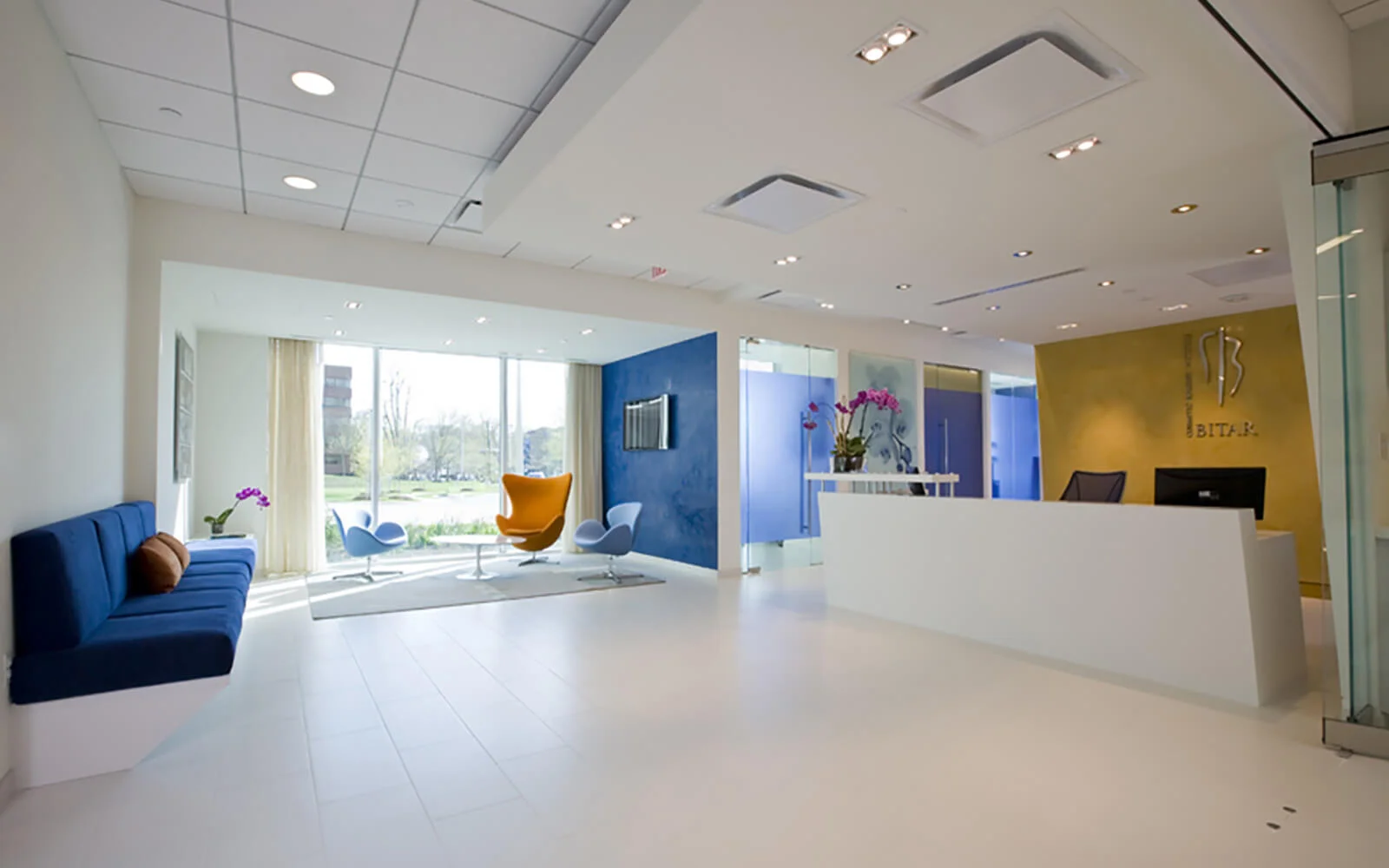 Interior view of a brightly lit, modern medical spa reception area with a white desk and floor. Features include a blue couch, an orange egg chair, and a yellow accent wall with the "DR. BITAR" logo.