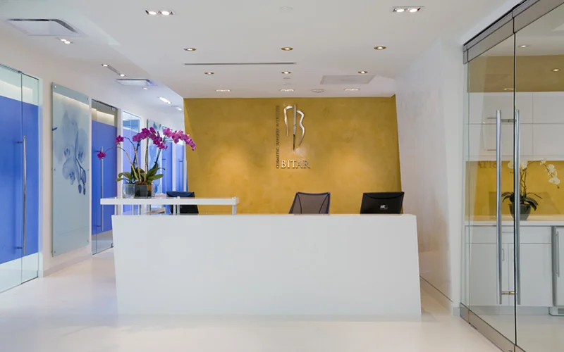 White reception desk and lobby area of a cosmetic surgery office with a gold-textured accent wall featuring a silver logo and the name Bitar Cosmetic Surgery Institute.