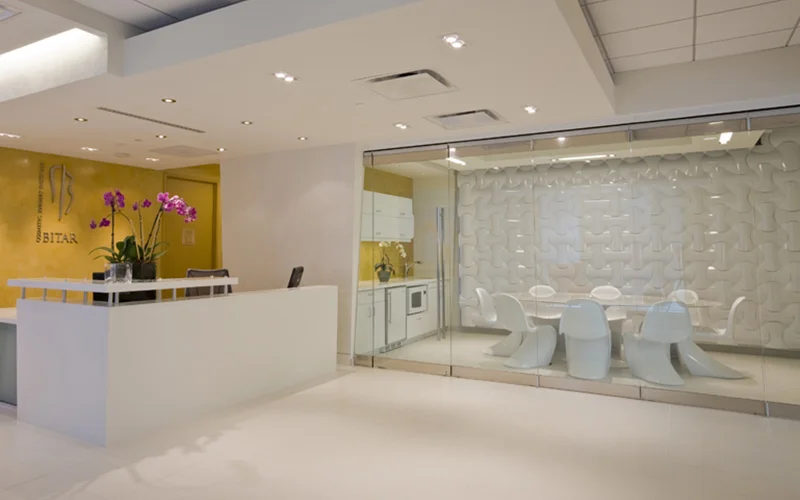 Interior view of a modern office space, showing a white reception desk with a vase of purple orchids. To the right, a glass-walled conference room features a white table and chairs against a textured white wall.