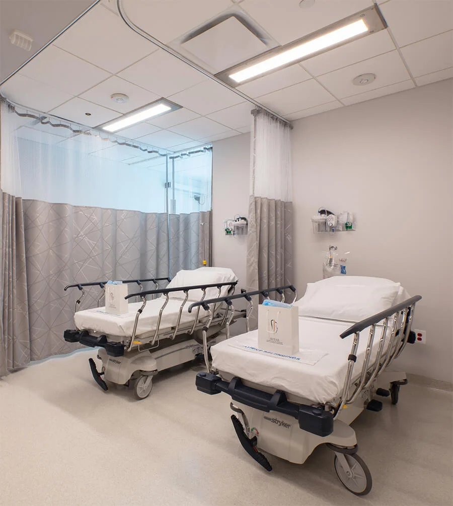 A clean, bright medical recovery room with two empty gurneys separated by curtains and packages from the Bitar Cosmetic Surgery Institute placed on the beds.