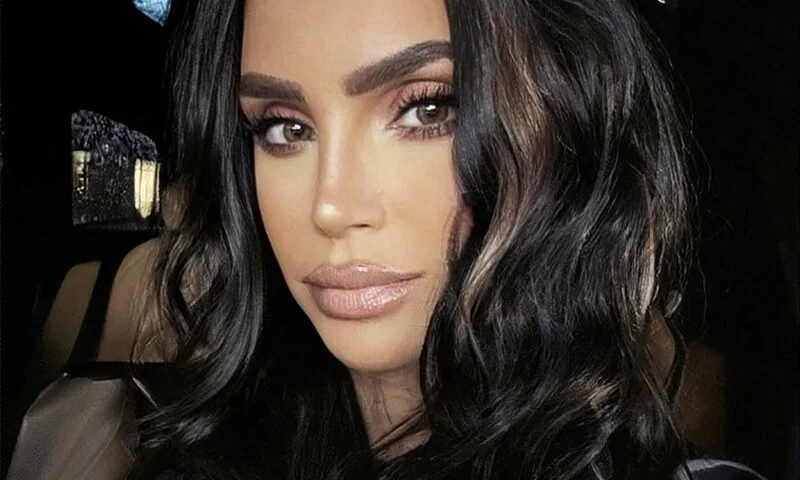 Close-up portrait of a woman with long dark wavy hair, brown eyes, and full lips, looking directly at the camera. She is wearing nude makeup.