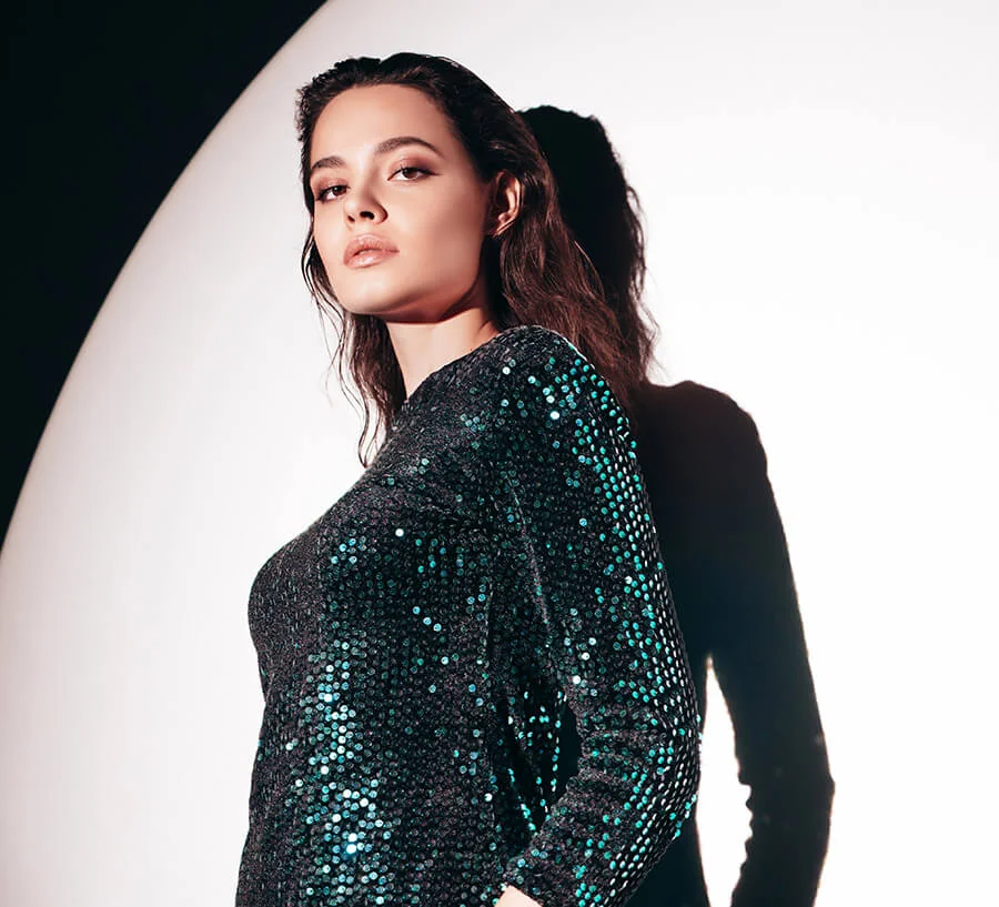 A striking portrait of a woman with wet-look hair and dark makeup, wearing a long-sleeved black dress covered in shimmering green sequins, casting a shadow on a white background.