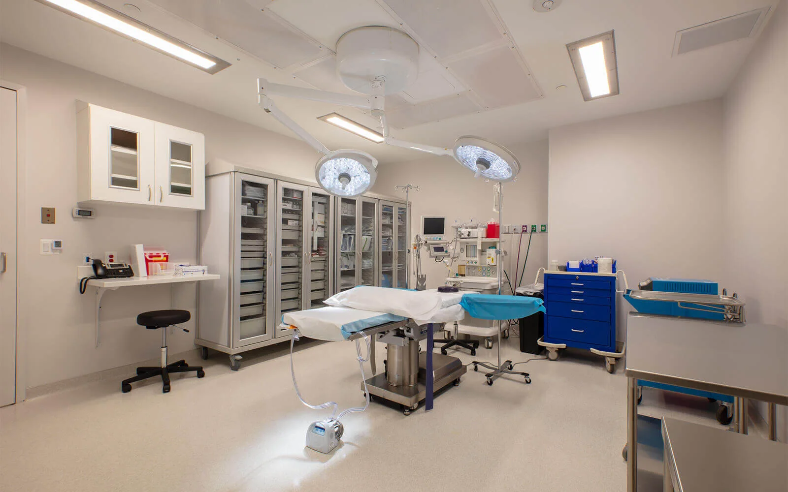 Operating room interior with a surgical table in the center, two overhead surgical lights, and various medical equipment and stainless steel cabinets lining the walls.