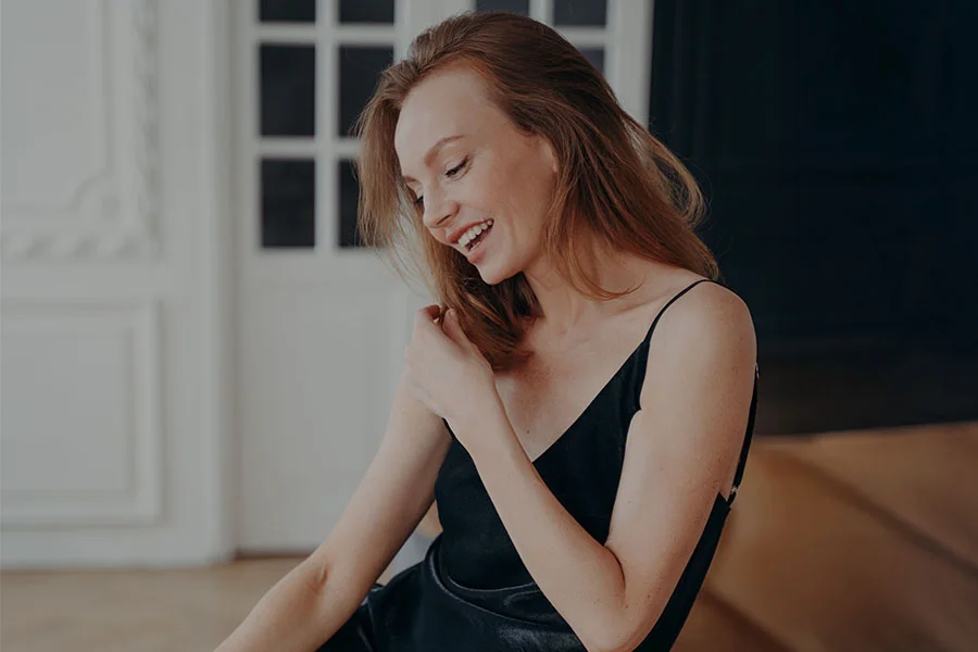 A light-skinned woman with shoulder-length reddish-brown hair is seated, smiling warmly and looking slightly to her left. She wears a dark, likely black, slip dress or top with spaghetti straps. Her joyful expression is complemented by soft, natural lighting. The background shows a light-colored wall and windows, creating a cozy, inviting atmosphere. - Arm Lift in Fairfax, PA