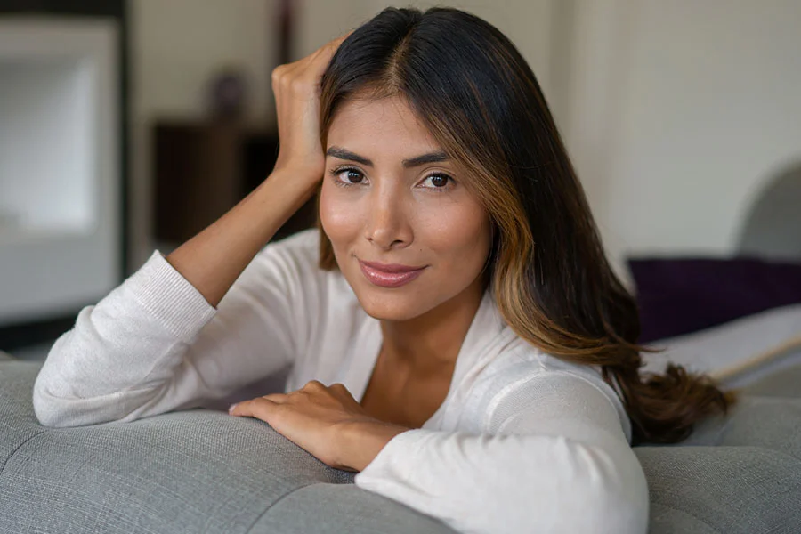 A woman with long dark brown hair and light highlights sits on a light gray couch, leaning back with a relaxed, gentle smile. She wears a cream long-sleeved top, resting one hand against her head. The blurred background suggests a cozy living space with muted gray and purple tones. - BBL Photo Rejuvenation in Fairfax, PA