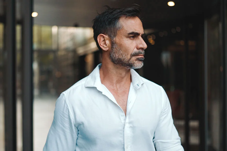 A mid-range close-up of a middle-aged man with short, dark graying hair and a neatly trimmed gray beard. He gazes off to his right with a neutral, thoughtful expression. He wears a pale off-white button-down shirt with the top buttons undone, slightly revealing his chest. The background is blurred, showing dark gray and black structures, possibly a modern building with glass walls, suggesting a business or urban setting. - Bio Identical Testosterone Pellet Treatment in Fairfax, PA