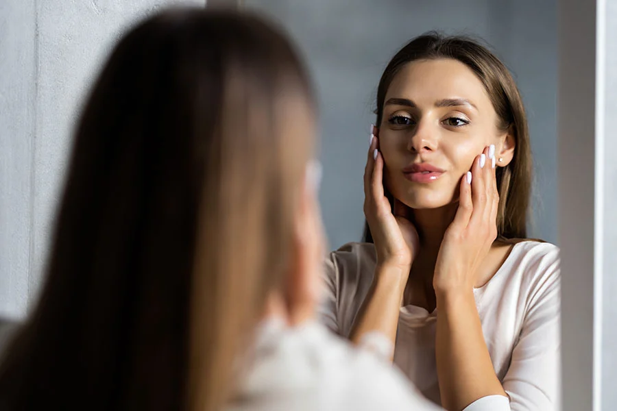 A woman wearing a light beige robe looks at her reflection in a mirror, gently touching her face and neck with a pleasant, focused expression. Her skin appears smooth, and a thin red bracelet is visible on her wrist. A blurred figure, likely her own, is seen in the reflection against a muted gray background. - Botox Cosmetics in Fairfax, PA