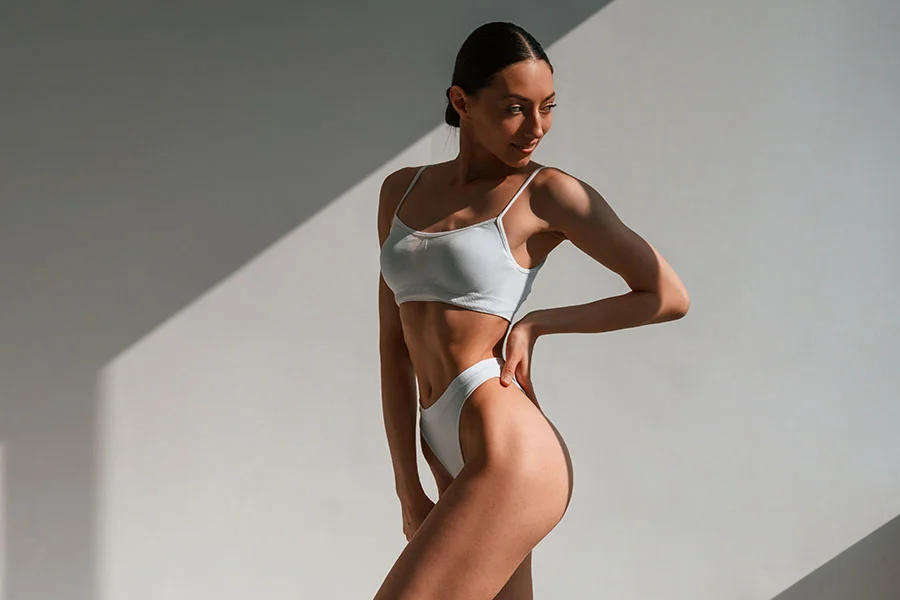A slender woman in a white sports bra and white underwear is standing with her hand on her hip, looking over her shoulder with a slight smile. The woman has her hair pulled back and is positioned in front of a white wall. The image features strong shadows, highlighting her silhouette and the contours of her body. - Brazillian Butt Lift in Fairfax, PA