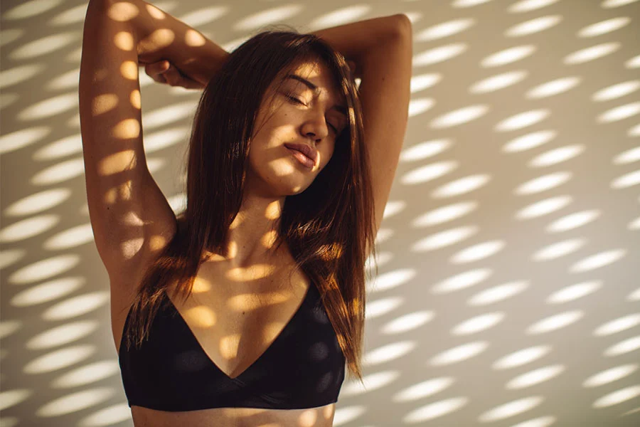 A young woman with long brown hair stands against a wall patterned with soft light and shadow. Her arms are raised above her head, and her eyes are closed in a calm, relaxed pose. She wears a simple black triangle bra. The dappled light creates an oval or dot-like pattern across the beige wall and her skin, adding depth and visual interest to the scene. - Breast Lift with Augmentation in Fairfax, PA