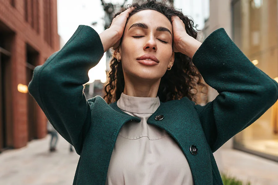 A woman with curly dark brown hair stands outdoors on a city street, wearing a dark teal-green coat over a light beige top. Her hands rest on her head, and her eyes are closed as she looks off into the distance. In the background are buildings, including one with reddish-brown brick and glass storefronts, and a paved street. - Brow Lift in Fairfax, PA