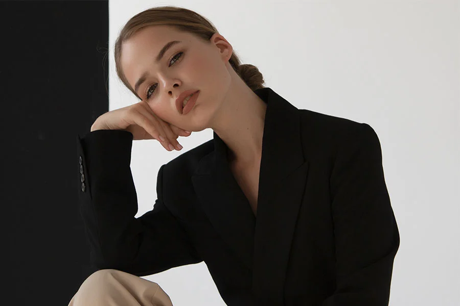 A woman in a black blazer and beige pants is posing for a photo. She has her hair pulled back in a bun and is resting her chin on her hand, looking directly at the camera with a serious expression. The background is a studio with a black panel on the left and a white wall on the right. The lighting is soft and even, highlighting her facial features. The image conveys a sense of sophistication and confidence. - Buccal Fat Removal in Fairfax, PA