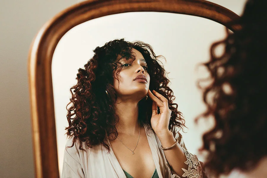 A young woman with long, curly brown hair, wearing a beige robe with lace trim. She is looking at her reflection in a mirror and touching her chin with her right hand. She is wearing a thin gold necklace and a beaded bracelet. - Chin Reduction Surgery in Fairfax, PA