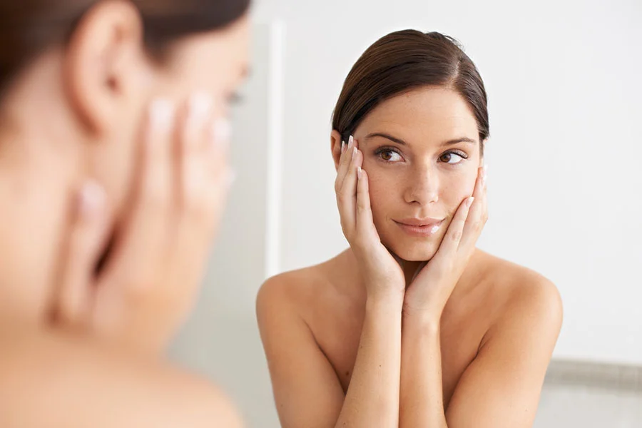 A woman with dark brown hair pulled back is seen in a bathroom mirror, her hands resting gently on her cheeks. Her expression is pleasant and thoughtful, with her shoulders and upper chest visible. Soft, diffused light illuminates her face and body. The background is a plain, light-colored wall, slightly blurred, with part of her reflection softly out of focus in the foreground. - Dysport in Fairfax, PA