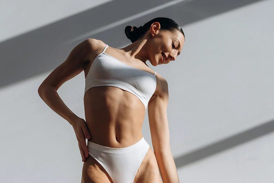 A woman is shown from the mid-section up in a three-quarter view, lit from above and the side. The light casts long, thin striped shadows across the light gray wall behind her. She wears a simple white lingerie set consisting of a supportive crop top and fitted briefs. Her posture is relaxed yet confident, with arms slightly bent and one hand resting near her waist. Her gaze is directed downward, expression soft, hair pulled back in a bun. Her smooth, even-toned skin and toned form are highlighted by the lighting. - Emsculpt Neo in Fairfax, PA