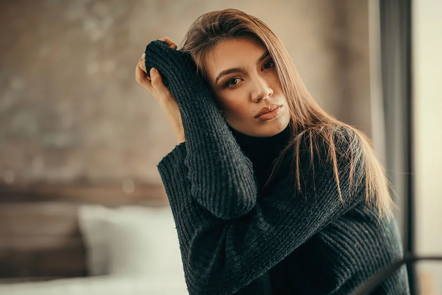 A serious-looking young woman with long, brown hair is posing while wearing a dark, ribbed turtleneck sweater. She is resting her chin on her hand, with her arms crossed and tucked into her sweater sleeves. Her gaze is directed at the camera with a confident expression. The background is a soft-focus, indoor setting with warm, natural light. - Eyelid Surgery in Fairfax, PA