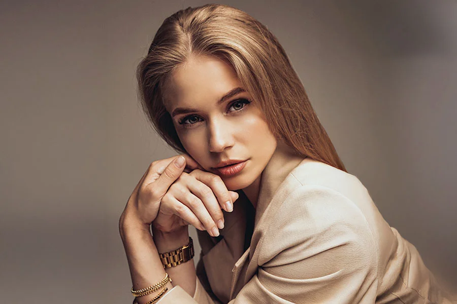 A young woman with long, blonde hair, wearing a beige blazer. She is seated, looking directly at the camera with her hands clasped together near her chin. She is wearing a gold watch, two gold bracelets, and a ring with a large black and gold design. The background is a solid, light gray. - Facial Fat Grafting in Fairfax, PA