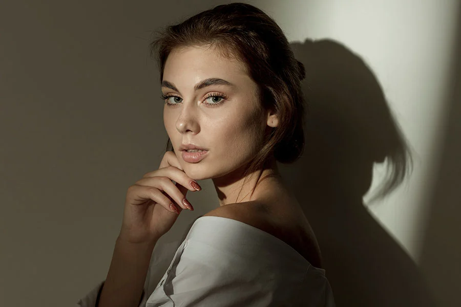 A woman with brown hair styled in a bun is shown from the mid-chest up in a three-quarter view, looking thoughtfully to her right with her hand resting gently on her chin. She wears a light-colored off-shoulder top. Soft, directional lighting casts a distinct shadow behind her on a neutral background, highlighting the contours of her face and neck, creating a sophisticated and elegant portrait. - Facial Treatments in Fairfax, PA