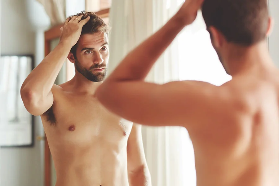 A shirtless man with a beard is looking at his reflection in a mirror. He is touching his hair with both hands, adjusting it. The man is looking directly at his reflection with a concerned expression. His chest has a noticeable prominence or fullness, which is a key detail in the context of the associated article about gynecomastia. The lighting is soft and natural, suggesting a bedroom or bathroom setting. The image captures a moment of self-scrutiny and body image concerns. - Gynecomastia in Fairfax, PA