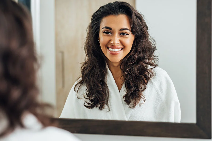 A woman with dark curly hair smiles brightly at her reflection in a mirror with a dark-brown wooden frame. She wears a white bathrobe, and her teeth are visible as she grins. A hint of another person’s dark curly hair appears in the reflection. The light-colored background suggests a bathroom or similar setting. - Hydrafacialt in Fairfax, PA