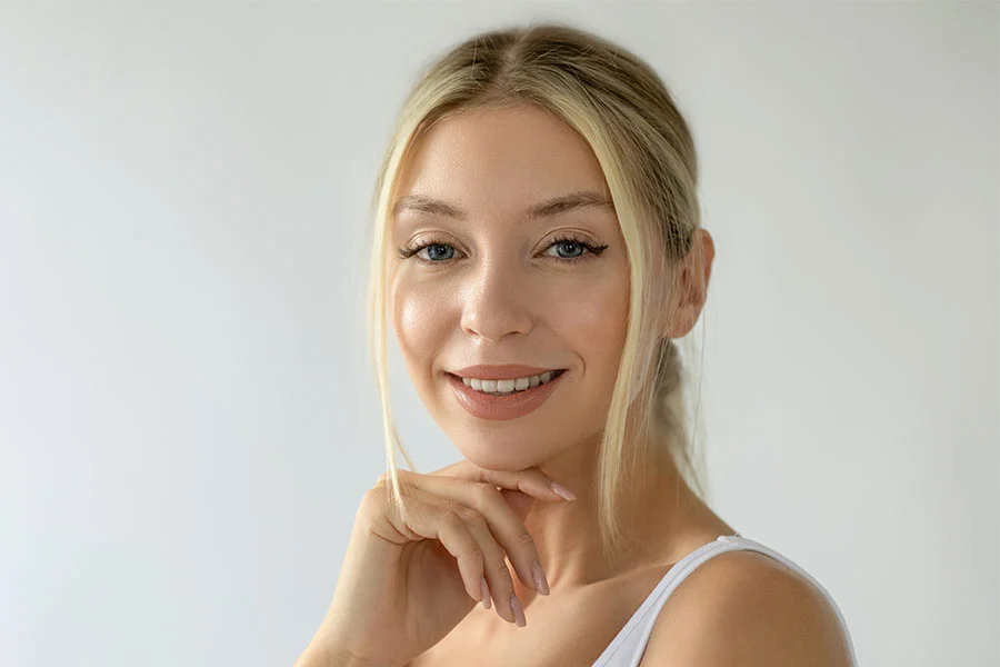 A woman with a short, black bob haircut, wearing a white spaghetti strap top. She is leaning on a white table with her arms crossed and her head tilted, looking directly at the camera. Her hair is slightly windswept, and she is wearing a thin silver earring. The background is a solid white. - IV Infusion Therapy in Fairfax, PA