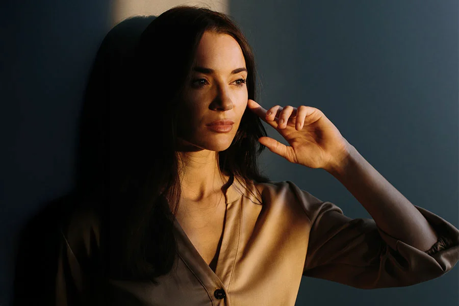 A portrait of a young woman with long, brown hair, wearing a white spaghetti strap top. She is looking away to her right with a pensive expression, and her hair is draped over her shoulders. The background is a solid gray. - Juvenderm Voluma XC in Fairfax, PA