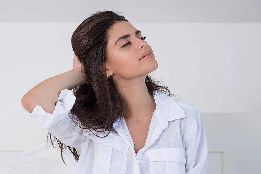 A beautiful Asian woman with short, black hair, wearing a white spaghetti strap top. She is seated, looking directly at the camera with her right hand on her left shoulder. The background is a solid white. - Kybella in Fairfax, PA