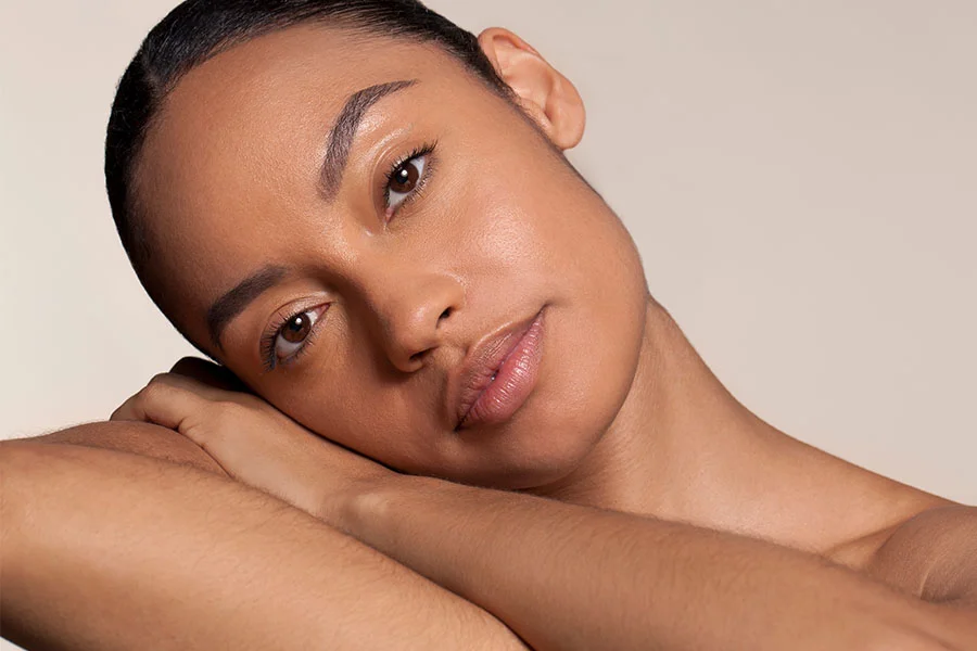 A young woman with a long, brown bob haircut, wearing a black slip dress with a lace neckline. She is seated on a sofa with pillows, looking directly at the camera with her hands clasped together in her lap. She is wearing a large gold cuff bracelet on her left wrist and a large gold earring on her right ear. A window with white blinds and a vase of dried plants are in the background. - Latisse in Fairfax, PA