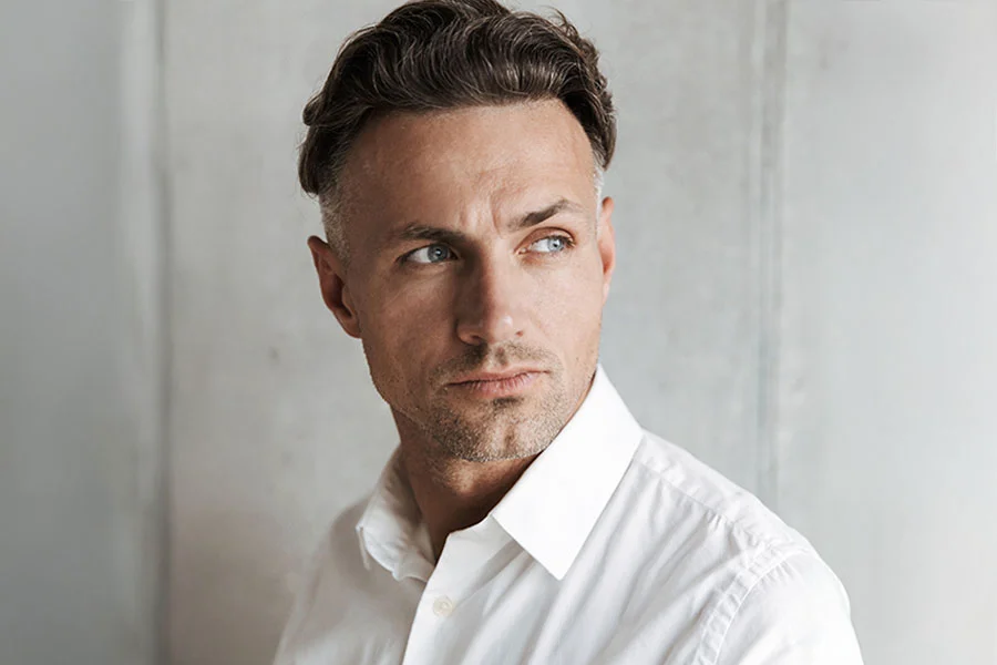 A man with dark and graying hair, wearing a white collared shirt, looking thoughtfully to his left. He has a serious expression on his face and is standing against a gray wall. - Male Eyelid Surgery in Fairfax, PA