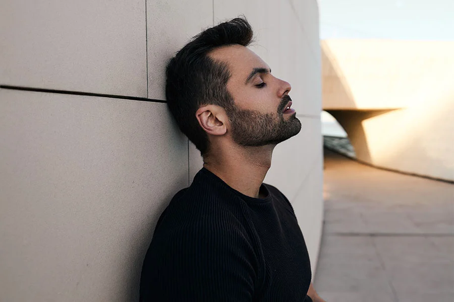 A head-and-shoulders shot of a man with a beard and dark hair, leaning his head back against a beige-colored wall. His eyes are closed, and his mouth is slightly open. He is wearing a black crewneck sweater. The background is a blurry outdoor scene with a modern building and a body of water. - Male Neck Lift in Fairfax, PA