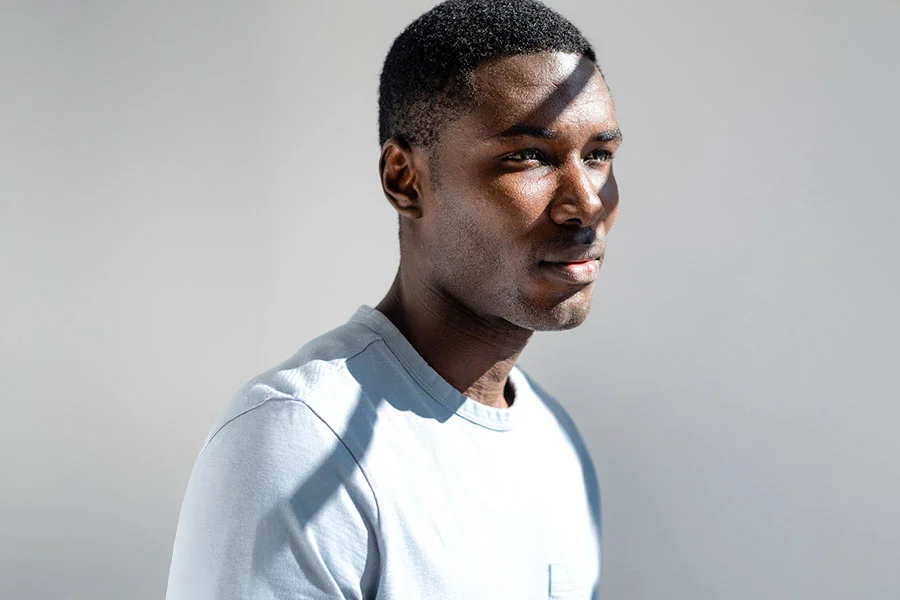 A head-and-shoulders shot of a Black man with short dark hair, wearing a light blue t-shirt. He is looking to the side, and a strong beam of sunlight creates a sharp diagonal line of light and shadow across his face. The background is a solid light gray color. - Male Rhinoplasty in Fairfax, PA