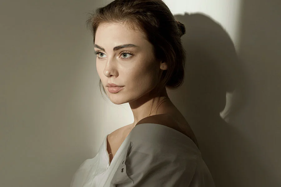 A woman with brown hair styled in a bun is shown from the mid-chest up in a three-quarter view, looking thoughtfully to her right with her hand resting gently on her chin. She wears a light-colored off-shoulder top. Soft, directional lighting casts a distinct shadow behind her on a neutral background, highlighting the contours of her face and neck, creating a sophisticated and elegant portrait. - Model Rhinoplasty in Fairfax, PA
