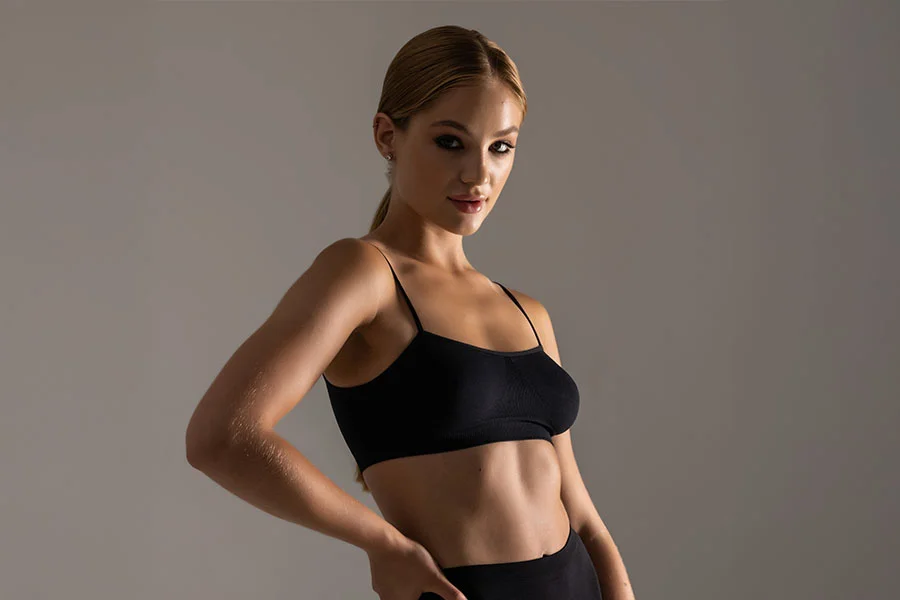 A woman with blonde hair tied in a ponytail is shown in a three-quarter shot, looking at the camera. She is wearing a black sports bra and appears to be in an athletic stance, with one hand on her hip. The background is a solid light gray color. - Morpheus8 in Fairfax, PA