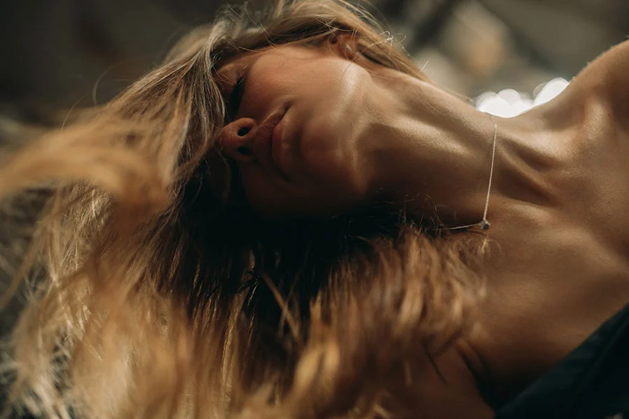 Close-up of a woman’s head and upper torso with flowing light brown hair, some strands falling over her face and shoulders. Her light tan skin appears smooth, and her head is tilted slightly back, revealing her neck. A thin silver necklace rests delicately around her neck. The background is dark and blurred, with warm lighting highlighting her features, creating a soft and delicate portrait. - Neck and Chin Liposuction in Fairfax, PA