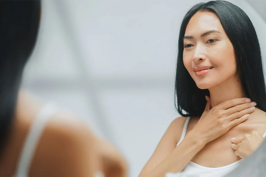 A young woman with long, straight black hair is shown in a mirror, looking at her reflection. She is smiling and touching the side of her neck with one hand. She is wearing a white tank top. The background is blurry and out of focus. - Neck Lift in Fairfax, PA