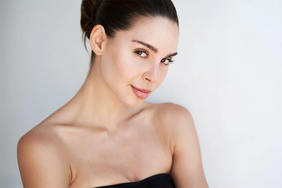 A close-up of a woman with her hair pulled back and her eyes closed, looking down and to her left. She has her left hand resting on her right shoulder. The background is a solid, light color. - Non-Surgical Facelift in Fairfax, PA