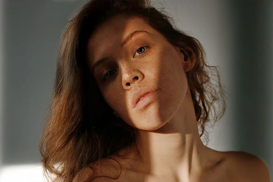 Close-up of a woman with shoulder-length, wavy dark brown hair and light tan skin with freckles across her nose and cheeks. Soft lighting casts shadows that highlight her features. She has light green eyes and natural rosy pink lips. The background is softly blurred, emphasizing her face and neck in a natural, gentle style. - Non-Surgical Rhinoplasty in Fairfax, PA