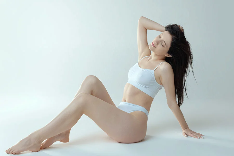 A young woman in a white sports bra and underwear is sitting on a white floor with a light gray background. She is leaning back on one arm and has her other hand in her dark hair, with her eyes closed and head tilted back slightly. Her legs are extended, one straight and one slightly bent. - Non-Surgical Tummy Tuck in Fairfax, PA