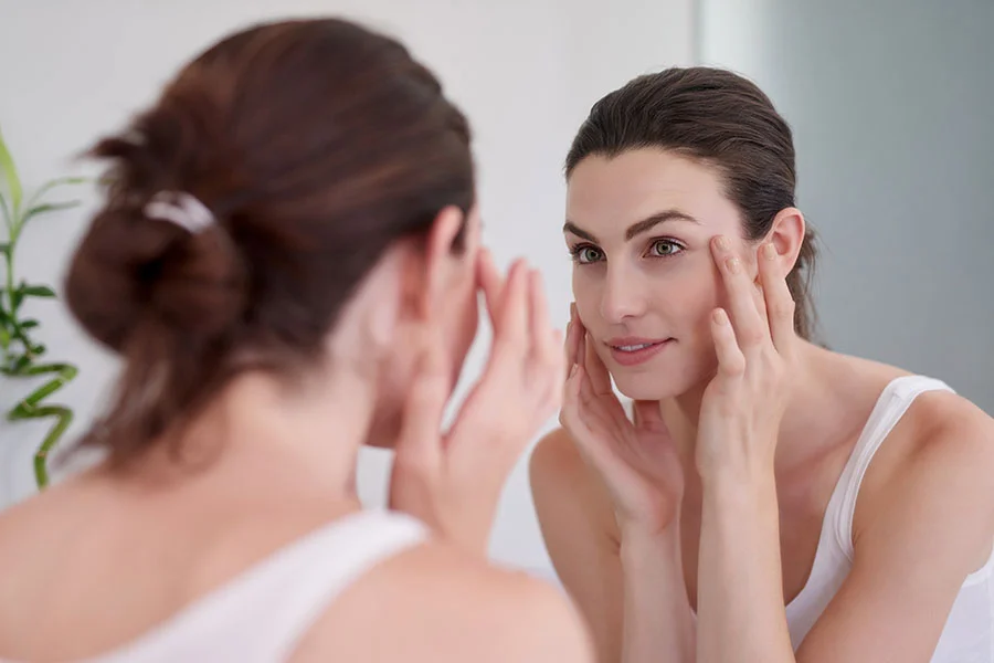 A young woman with long, dark hair looks intently at her reflection in a mirror, her face framed by her hands as she gently touches the skin around her eyes, possibly examining or applying a product. She is wearing a simple white tank top, and the background is a soft, blurred reflection, creating a sense of intimacy and focus on her skincare routine. - Plasma Pen in Fairfax, PA