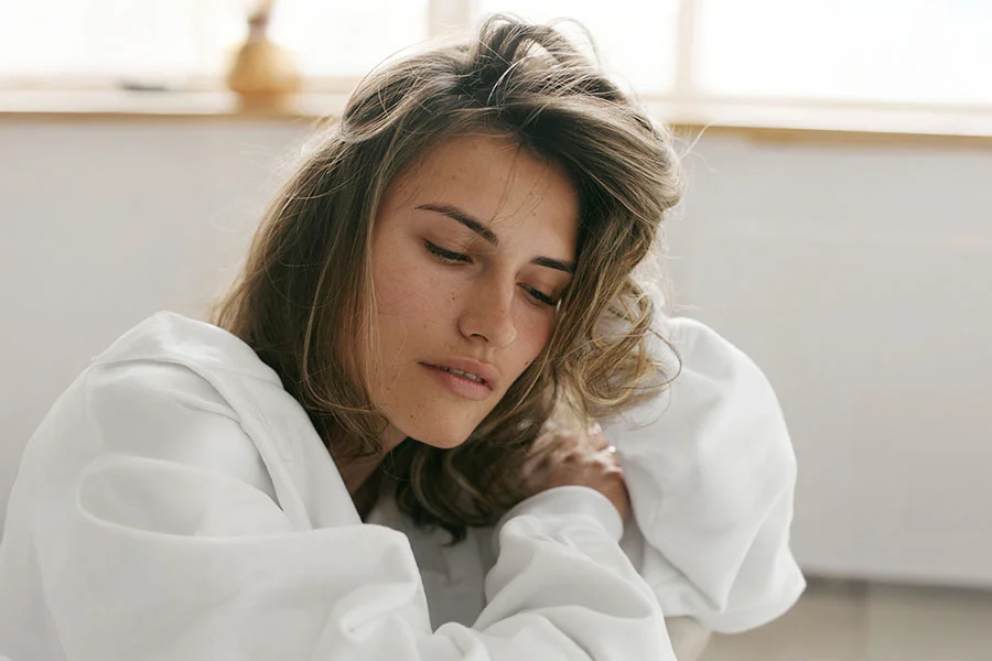 Close-up of a young woman with light brown, slightly tousled hair framing her face. Her expression is neutral and contemplative, with her gaze directed slightly off to the viewer’s right. She has smooth, light-toned skin and wears a white, loose-fitting top. Soft, diffused lighting creates a gentle glow around her, with a window or light source visible in the bright, airy background. - Restalyne in Fairfax, PA