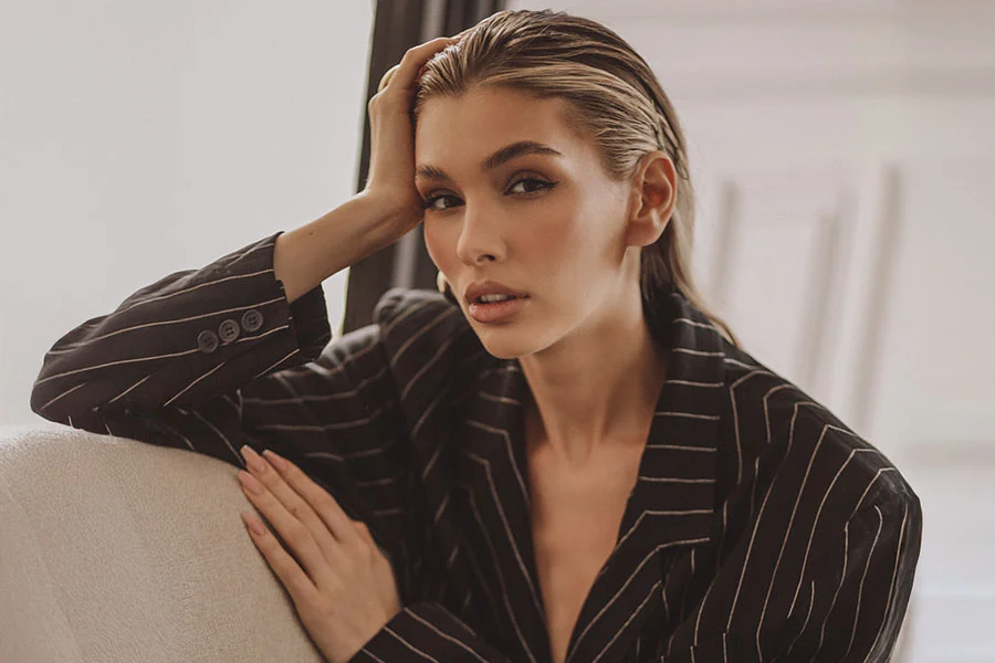 A young woman with slicked-back, wet-look hair is shown in a three-quarter shot. She is wearing a black pinstripe blazer with buttoned cuffs and is leaning on a light-colored couch. She is looking directly at the camera with a neutral expression. - Revision Rhinoplasty in Fairfax, PA