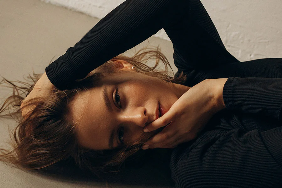 A young woman with light brown hair, wearing a black long-sleeved shirt, lying on her side on a gray surface with her head resting on her arms and looking directly at the camera. - Rhinoplasty Reduction in Fairfax, PA