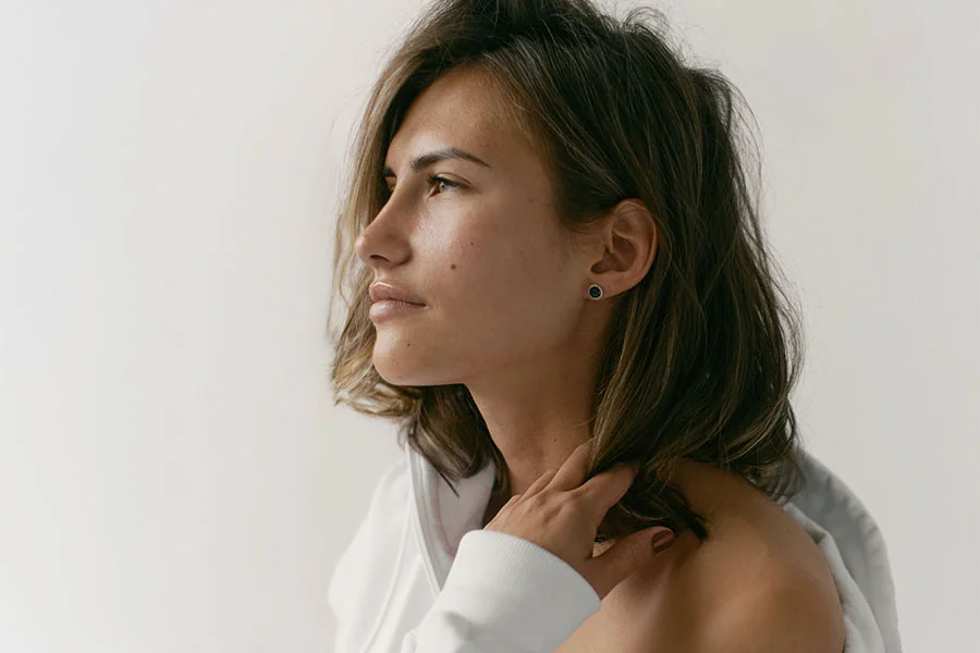 A young woman with brown, shoulder-length hair looks to the left, with her profile visible. She is wearing a white hoodie or top that is pulled down to expose her shoulder, and her hand is resting near her neck, gently touching her hair. She has a dark stud earring in her ear. The background is a plain, light color. - Ultrasonic Rhinoplasty in Fairfax, PA